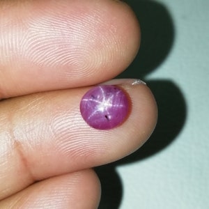 Könnte beinhalten: Ein runder, polierter Rubin-Edelstein im Cabochon-Schliff. Der Rubin hat eine tiefrosa-rote Farbe mit einem Sechs-Strahl-Stern-Effekt. Der Stern ist weiß und scheint auf der Oberfläche des Steins zu schweben. Der Edelstein wird zwischen zwei Fingern gehalten.