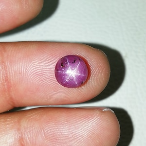 Könnte beinhalten: Ein polierter, runder Sternrubin-Edelstein. Der Edelstein hat eine tiefrosa-rote Farbe mit einem sechsstrahligen Sterneffekt auf der Oberfläche. Der Edelstein wird zwischen zwei Fingern gehalten.
