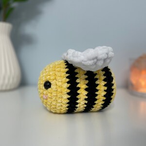 May include: A crocheted bee plush toy with yellow and black stripes and a white fuzzy wing. The bee has a black eye and a pink nose.