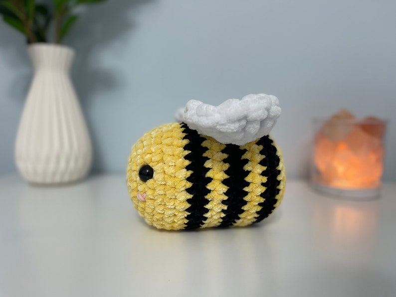 May include: A crocheted bee plush toy with yellow and black stripes and a white fuzzy wing. The bee has a black eye and a pink nose.