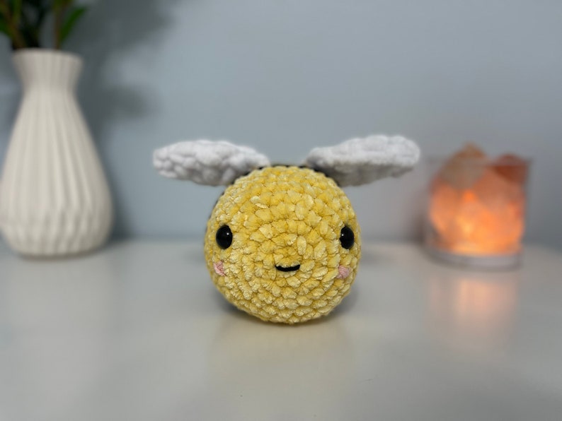 May include: A crocheted plush bee toy with a yellow and white fuzzy body, black eyes, and a pink smile. The bee has white wings.