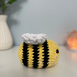 May include: A crocheted bee plush toy with a yellow and black striped body and a white fuzzy head. The bee has black eyes and a pink mouth.