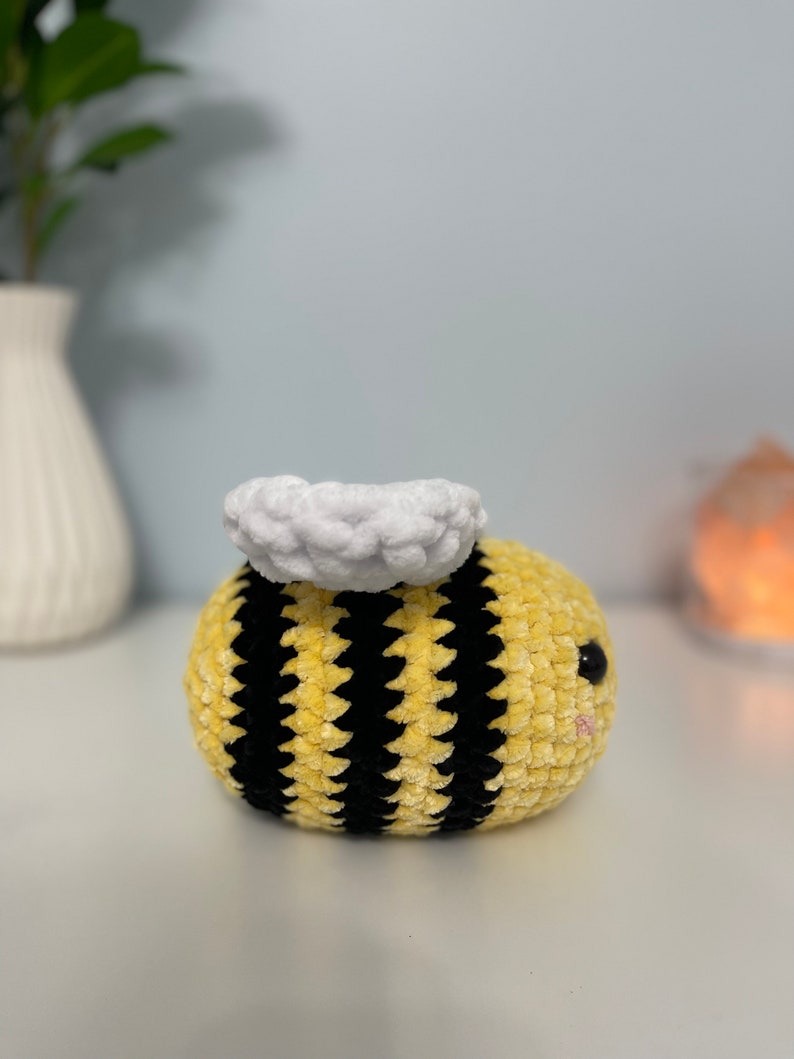 May include: A crocheted bee plush toy with a yellow and black striped body and a white fuzzy head. The bee has black eyes and a pink mouth.