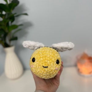 May include: A crocheted plush bee toy with a yellow body, white wings, and black eyes. The bee has a smiling face with pink cheeks.