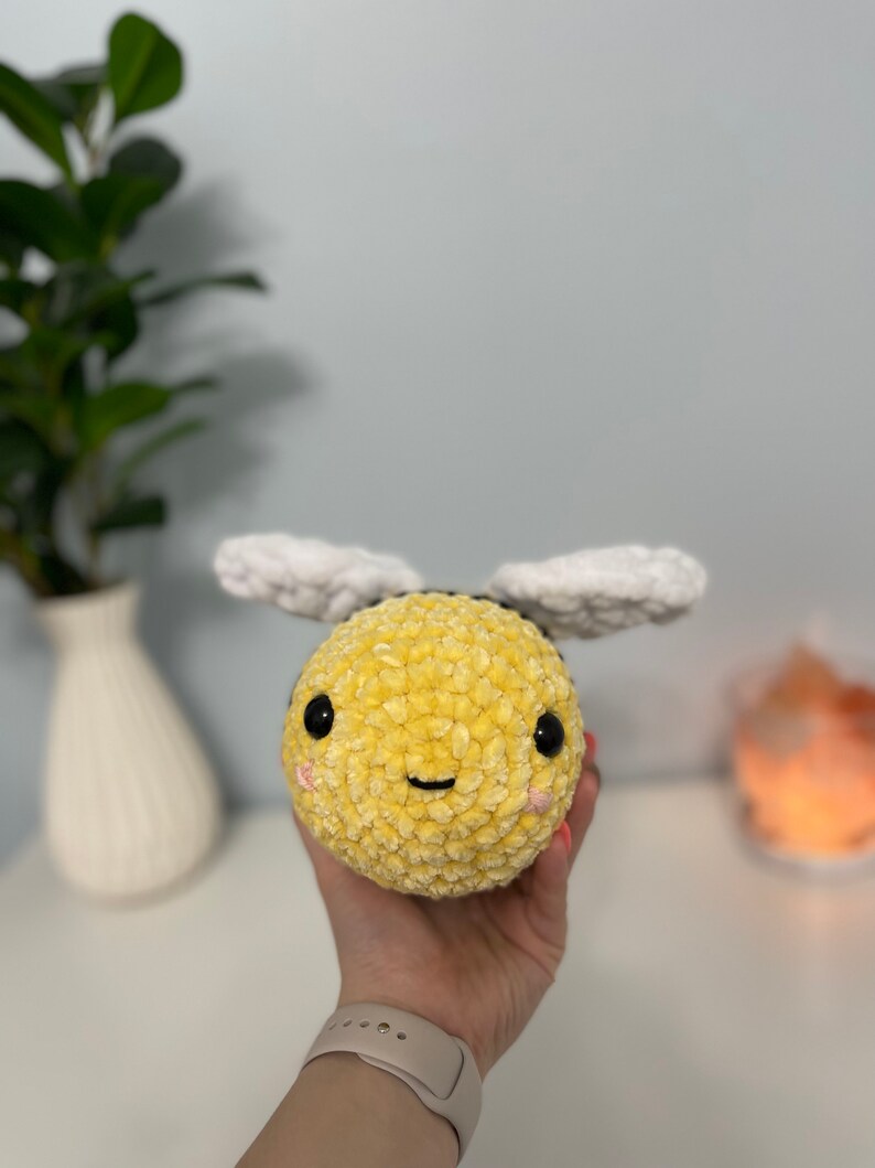 May include: A crocheted plush bee toy with a yellow body, white wings, and black eyes. The bee has a smiling face with pink cheeks.