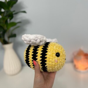 May include: A crocheted plush bee toy with yellow and black stripes, white wings, and black eyes. The bee is being held in a hand.