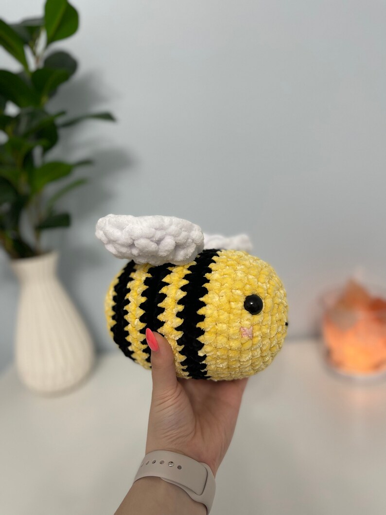 May include: A crocheted plush bee toy with yellow and black stripes, white wings, and black eyes. The bee is being held in a hand.