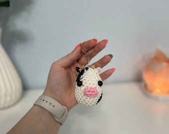 May include: A black and white cow keychain made of rubber bands. The cow has a pink nose and black eyes. The keychain is attached to a silver ring.