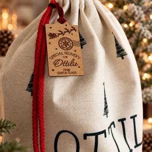 K&ouml;nnte beinhalten: Ein beigefarbener Geschenksack mit Kordelzug, bedruckt mit schwarzen Tannenb&auml;umen und dem Namen Ottilie. Ein Holzanh&auml;nger an einer roten Kordel zeigt einen North Pole Post Office Stempel, eine Silhouette von Santas Schlitten und den Text Special Delivery for Ottilie from Santa Claus.