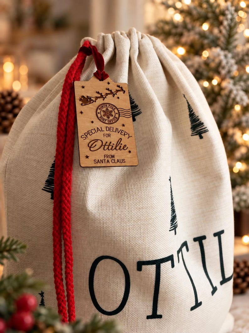 K&ouml;nnte beinhalten: Ein beigefarbener Geschenksack mit Kordelzug, bedruckt mit schwarzen Tannenb&auml;umen und dem Namen Ottilie. Ein Holzanh&auml;nger an einer roten Kordel zeigt einen North Pole Post Office Stempel, eine Silhouette von Santas Schlitten und den Text Special Delivery for Ottilie from Santa Claus.