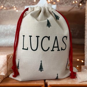 K&ouml;nnte beinhalten: Ein beigefarbener Geschenksack mit Kordelzug aus strukturiertem Stoff, bedruckt mit dem Namen LUCAS in gro&szlig;en schwarzen Gro&szlig;buchstaben. Der Sack ist mit kleinen schwarzen Tannenbaum-Illustrationen verziert und hat rote Kordeln.
