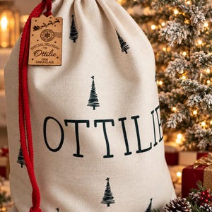 K&ouml;nnte beinhalten: Ein beigefarbener Geschenksack mit Kordelzug, bedruckt mit einem schwarzen Muster aus Tannenb&auml;umen und dem Namen OTTILIE in gro&szlig;en Buchstaben. Ein roter Kordelzug verschlie&szlig;t den Sack, und ein Holzanh&auml;nger tr&auml;gt die Aufschrift SPECIAL DELIVERY FOR OTTILIE FROM SANTA CLAUS.