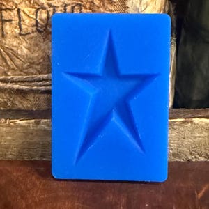 May include: A rectangular blue silicone mold featuring a single, centered five-pointed star indentation. The mold has a smooth, flat surface surrounding the star shape, which is recessed into the material, creating a clear and defined star outline.