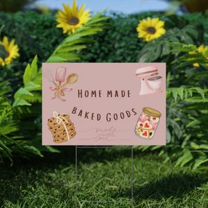 May include: A pink sign with the words "HOME MADE BAKED GOODS" and illustrations of baking tools, cookies, and a jar of heart-shaped treats. The sign is displayed outdoors in a grassy area.