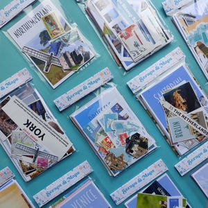 May include: A collection of travel-themed scrapbooking kits, each packaged in a clear plastic sleeve. The kits feature various locations, including "Northumberland," "York," and "Venice," with images of landmarks and travel ephemera. The kits are arranged on a turquoise surface.