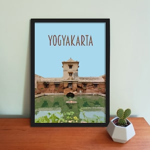 Pode incluir: Uma impressão emoldurada com uma moldura preta e um fundo azul claro. A impressão apresenta um edifício castanho com uma piscina verde na frente. O texto "YOGYAKARTA" está impresso em castanho na parte superior da impressão.