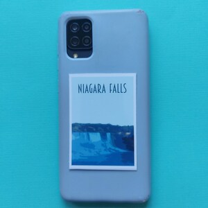 Może przedstawiać: Szare etui na telefon z naklejką przedstawiającą wodospad Niagara. Naklejka zawiera niebieski i biały obraz wodospadu z napisem "NIAGARA FALLS" w kolorze czarnym.
