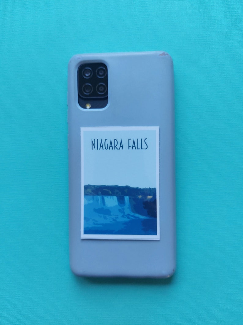 Może przedstawiać: Szare etui na telefon z naklejką przedstawiającą wodospad Niagara. Naklejka zawiera niebieski i biały obraz wodospadu z napisem "NIAGARA FALLS" w kolorze czarnym.