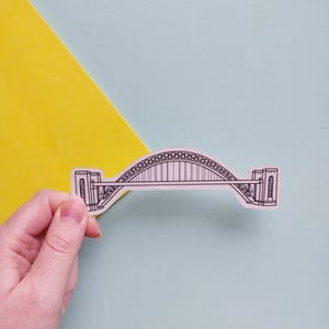 May include: A sticker depicting a black and white line drawing of a bridge. The bridge has an arched design with detailed support structures. The sticker is held against a backdrop of yellow and light blue.