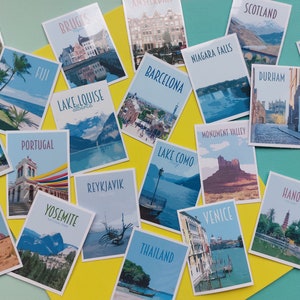Op de afbeelding: Een verzameling kleurrijke ansichtkaarten met verschillende reisbestemmingen over de hele wereld. De ansichtkaarten zijn gerangschikt op een geel en turquoise oppervlak. Enkele van de bestemmingen zijn: Schotland, Melbourne, Durham, Nieuw-Zeeland, Londen, Portugal, Lake Louise, Yosemite, Reykjavik, Lake Como, Veneti&euml;, Hanoi, Thailand, Brugge, Cotswolds, Singapore, Australi&euml;, Fuji, Barcelona, Niagara Falls en Monument Valley.