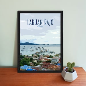 May include: A framed print with a colorful illustration of a town on a coast with a body of water and mountains in the background. The text "Labuan Bajo Flores" is at the top of the print.