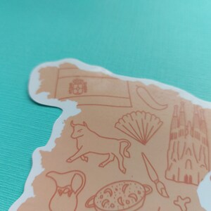 Peut inclure: Un autocollant repr&eacute;sentant l'Espagne avec diverses illustrations de la culture espagnole, notamment un taureau, un &eacute;ventail, une po&ecirc;le &agrave; paella, une &eacute;glise et le drapeau espagnol. L'autocollant est sur un fond turquoise.