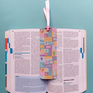 May include: A colourful bookmark with a pink, yellow and blue design. The bookmark features a variety of illustrations, including a moon, stars, a house, a tree and a road. The bookmark is perfect for adding a touch of whimsy to any book.