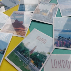 Op de afbeelding: Een verzameling vintage reisposters in postkaartstijl met verschillende bestemmingen, waaronder Monument Valley, Veneti&euml;, Hanoi, Nieuw-Zeeland, Londen en Durham. De posters zijn gerangschikt op een gele en turquoise ondergrond.