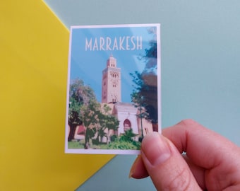 May include: A postcard with a photo of a tall, pink minaret with a blue sky and green trees in the background. The text "MARRAKESH" is printed in white at the top of the postcard.