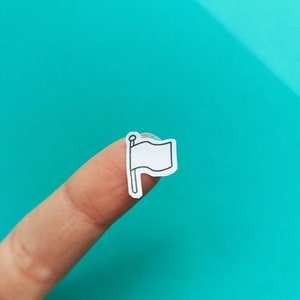 May include: A finger holding a white flag sticker with a black outline. The flag is on a stick and is waving in the wind.