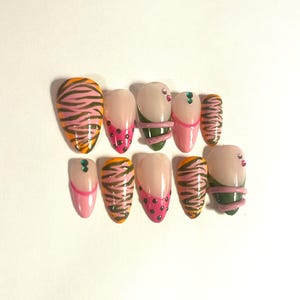 May include: A set of ten artificial nails in various designs. Some nails feature a tiger stripe pattern in orange, brown, and pink. Others have pink and black polka dots, or a pink and green swirl design. Some nails have small, decorative gems.
