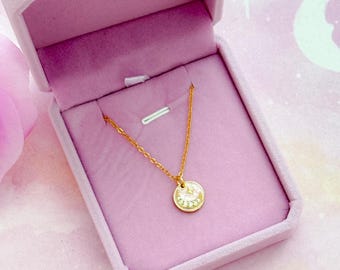 May include: A gold-colored necklace with a circular pendant featuring a clock design, displayed in an open, light pink velvet jewelry box. The box is set on a pink background with a floral and celestial theme, including a crescent moon and stars. Pink roses are also present.