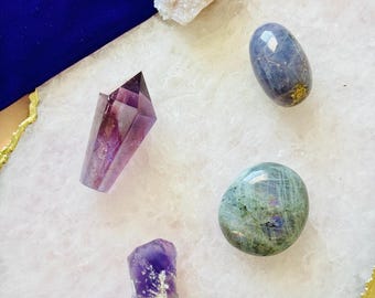 May include: An assortment of crystals and gemstones in various shapes and colors. Includes a large, faceted amethyst crystal, a tumbled blue-grey stone, and a cluster of small, white crystals. A blue velvet pouch is visible in the background.