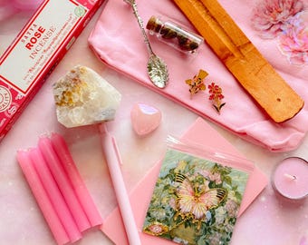 May include: A collection of pink items, including incense, candles, a pen, a heart-shaped crystal, and a butterfly-themed pin. A small glass bottle with a cork and a decorative spoon are also present. The items are arranged on a pink surface.