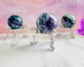 May include: Three polished crystal spheres in shades of blue, green, and purple, each displayed on an ornate silver-colored stand. The stands have a decorative, scrolled design. The background is a soft gradient of pink and purple.