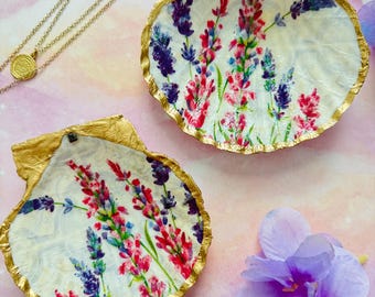 May include: Two seashell trinket dishes with gold leaf edges. The interior of each shell is painted with lavender flowers in shades of purple, pink, and green. A gold necklace and a purple flower are also visible.