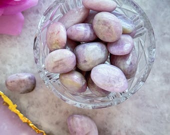 May include: A clear, cut-glass bowl filled with smooth, oval-shaped, light purple stones. The stones are a soft lavender color with subtle variations. The bowl sits on a light purple surface with a gold-edged border, and a pink rose is visible in the background.