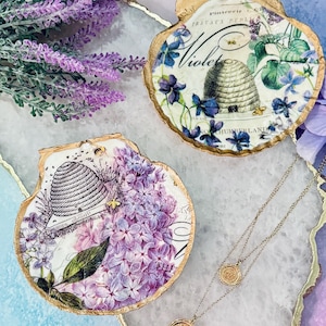 May include: Two decorative seashell trinket dishes with gold edges. One features a beehive and lilac design, the other a beehive and violet design. A delicate necklace with two round pendants is also visible.