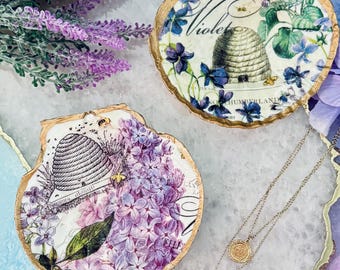 May include: Two decorative seashell trinket dishes with gold edges. One features a beehive and lilac design, the other a beehive and violet design. A delicate necklace with two round pendants is also visible.