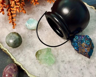 May include: A black cauldron with a handle, surrounded by various colorful crystals and gemstones. The crystals include a light blue, green, purple, and a blue-gold iridescent stone. The arrangement is set on a white surface with a gold trim.
