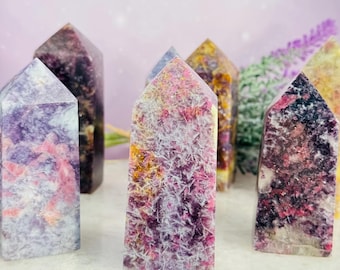 May include: A collection of colorful crystal towers. The crystals display a variety of colors, including pink, purple, yellow, and white. The crystals are arranged on a white and gold tray, with a soft purple background.