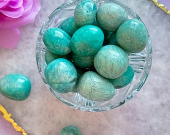 May include: A clear glass bowl filled with polished, oval-shaped turquoise stones. The stones have a mottled appearance and are scattered on a light purple surface with a gold trim. A pink rose is in the background.