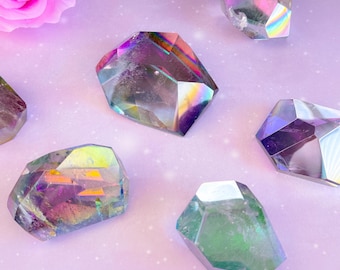 May include: Several iridescent, faceted crystals are scattered on a light purple surface. The crystals display a range of colors, including pink, green, and blue, with a rainbow effect. A pink rose is in the upper left corner.