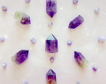 May include: Amethyst crystals in various shapes and sizes, arranged on a light pink surface. The crystals range in color from deep purple to pale lavender and are surrounded by smaller, iridescent stones. The arrangement creates a visually appealing display.