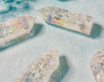 May include: Several clear quartz crystal points with iridescent rainbow hues are arranged on a light blue surface. The crystals are rectangular with a pointed top. Some blue flowers are in the background.