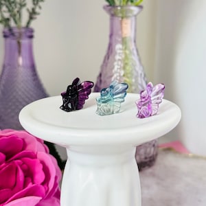 May include: Three small, carved crystal fairy figurines in shades of purple and blue sit on a white pedestal. The fairies have detailed wings and are displayed in front of purple vases with flowers.