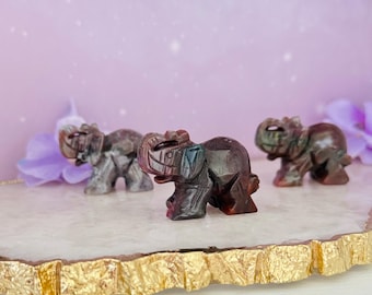 May include: Three carved stone elephant figurines in shades of brown, gray, and red are displayed on a gold-edged, white surface. The elephants are intricately detailed, showcasing their trunks, ears, and bodies. The background is a soft purple.