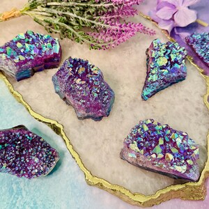 May include: An assortment of iridescent, purple, and blue aura quartz crystals. The crystals are arranged on a white and gold-edged tray, with sprigs of lavender and a purple flower in the background. The crystals have a sparkling, textured surface.
