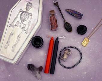 May include: A collection of items including a clear coffin-shaped box with a skeleton design, a small glass vial with red contents, a decorative metal spoon with a crystal handle, a raw iridescent crystal, a black rose, a gold-toned rectangular pendant necklace, a black candle holder, one orange and one black candle, a small mushroom figurine, a beaded bracelet, and a dark crystal cluster.