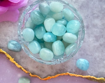 May include: A clear glass bowl filled with smooth, tumbled aquamarine stones. The stones vary in shades of blue and green. A pink rose and gold-edged purple surface provide the backdrop.